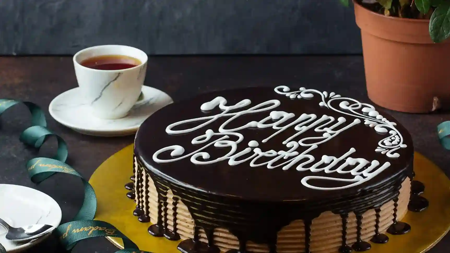 Chocolate birthday cake with Happy Birthday icing, tea cup, and plant on table