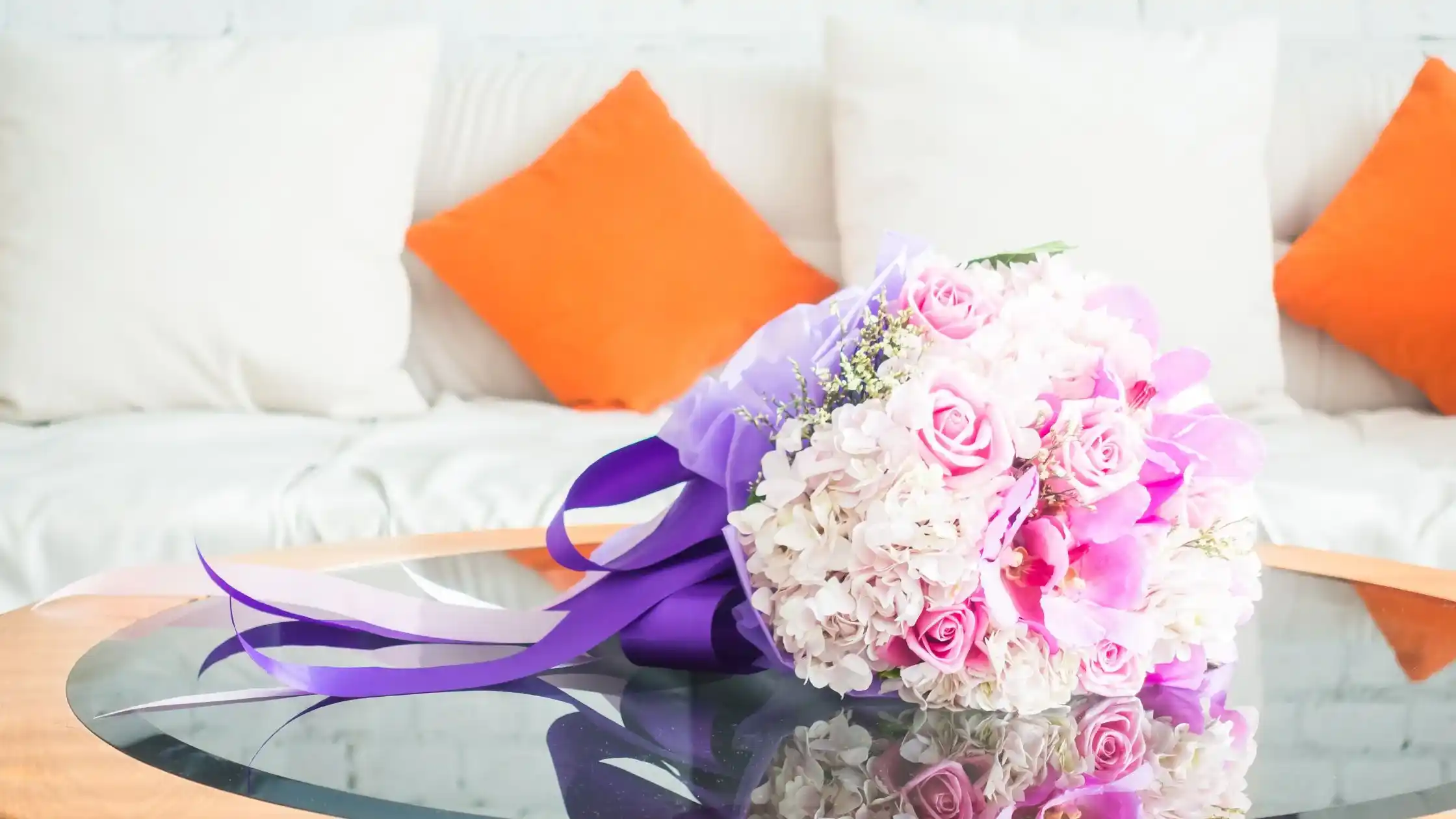 Elegant pink and white flower bouquet wrapped in purple ribbon placed on a glass table in a modern room with white sofa and orange cushions.